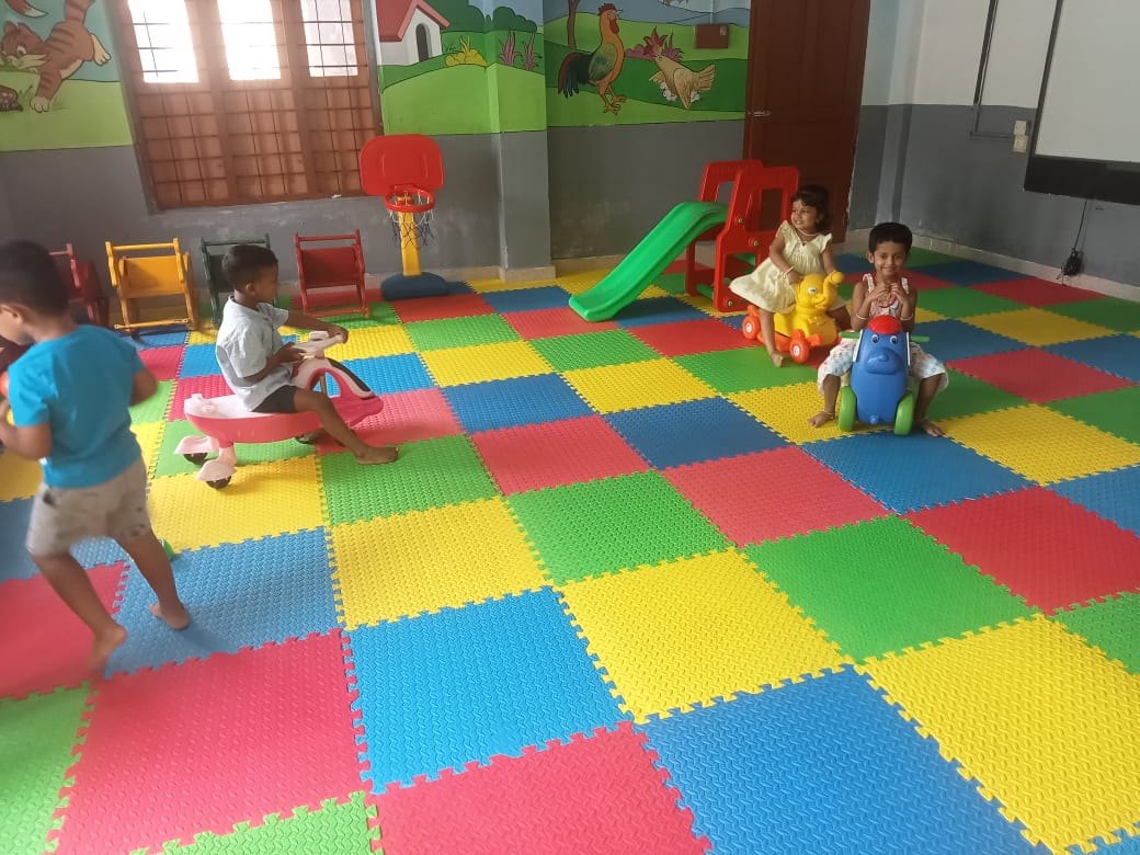 Pre-primary children riding toy cars and playing on a slide on a colourful foam-tiled floor, with animals painted on the walls